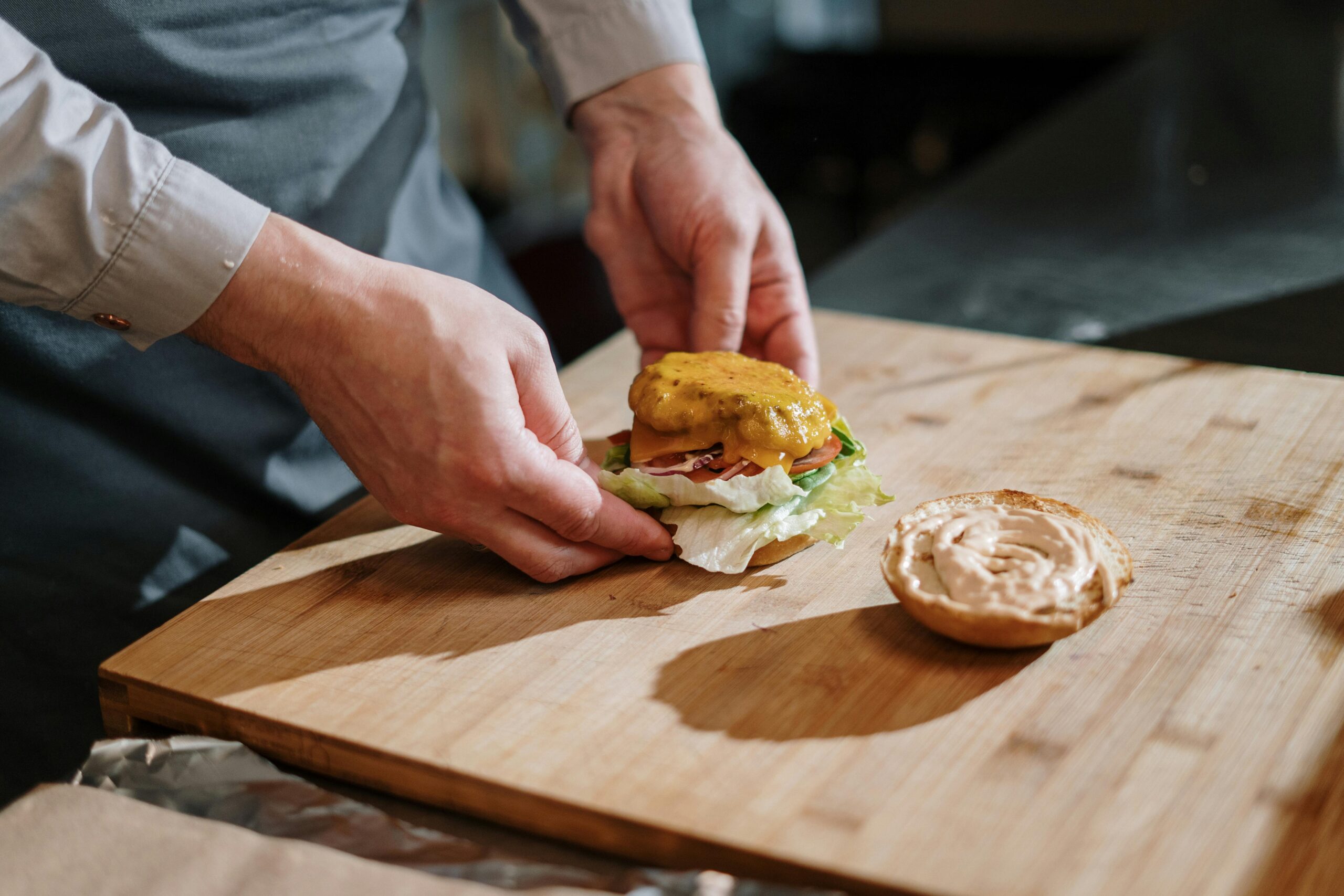 Chef crafting a delicious cheeseburger with lettuce, tomato, and cheese on a wooden cutting board indoors.